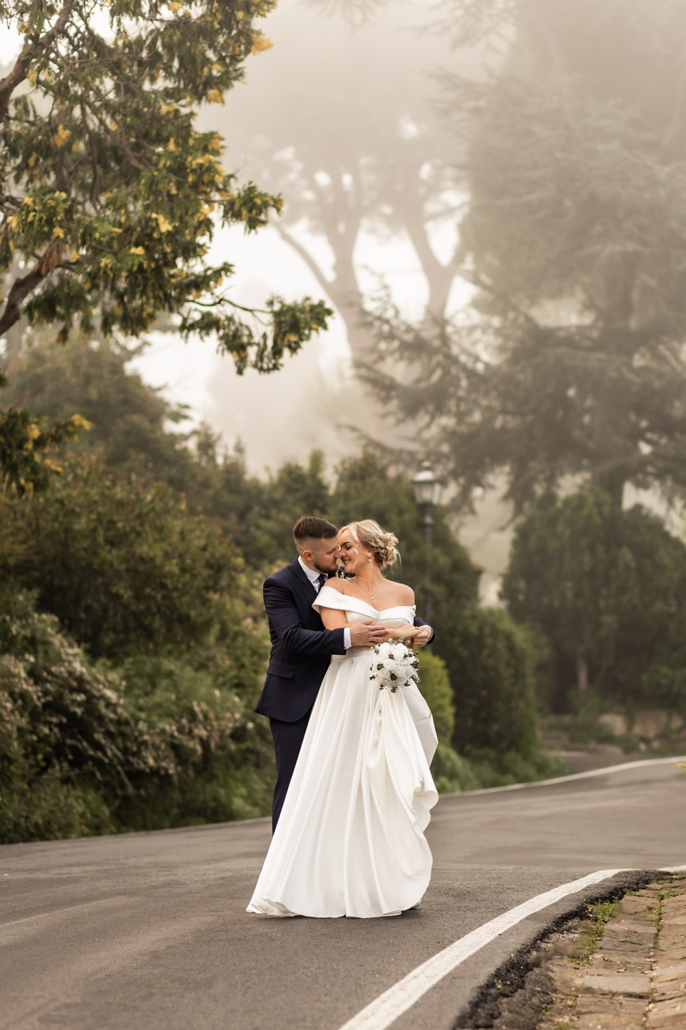 Couple standing on a foggy street early in the morning cuddling in their wedding gown.