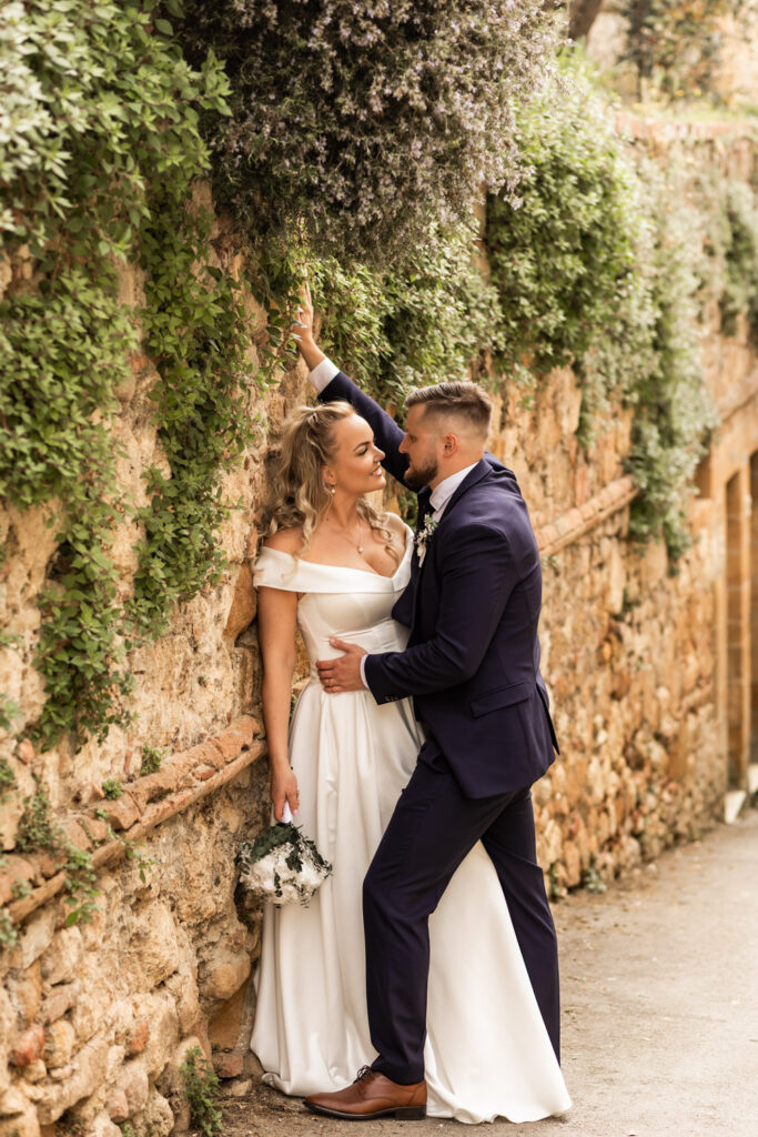 Husband kissing wife's forehead while leaning against a plant-covered wall in a small Italian town.