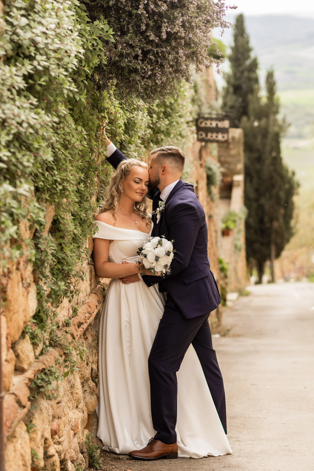 Husband kissing wife's forehead while leaning against a plant-covered wall in a small Italian town.