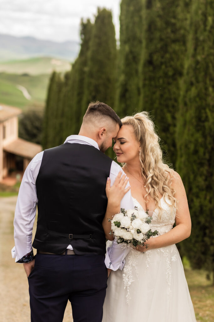 Husband and wife having a sweet moment in their wedding gown. They are standing in front of cypress trees in Tuscany.