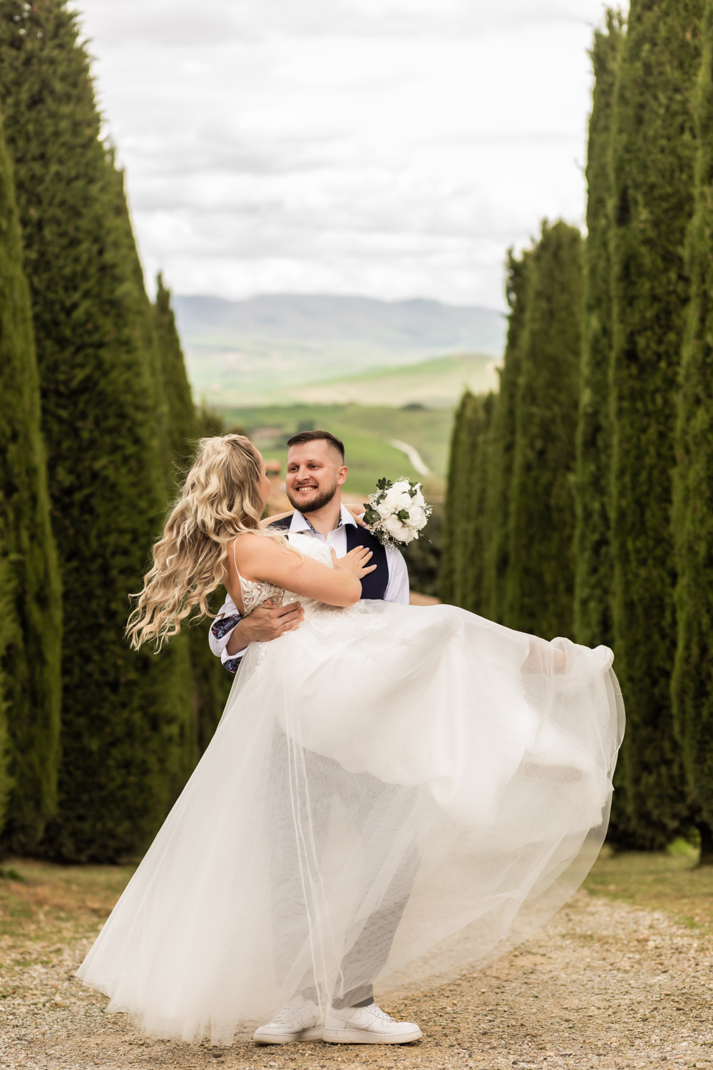 Husband holding wife in his hands in their wedding gown. They are standing in front of cypress trees in Tuscany.
